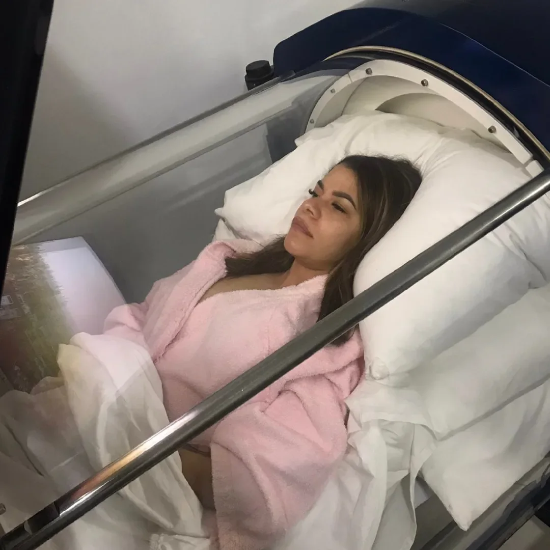Patient in a pink robe relaxing inside the clear Bella Oxygen hyperbaric chamber